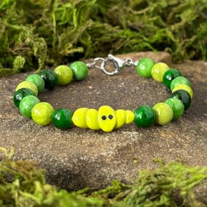 May include: A beaded bracelet featuring a mix of green and yellow beads. The bracelet has a silver clasp and a central yellow bead with a face design. The beads vary in shades of green, creating a vibrant accessory.