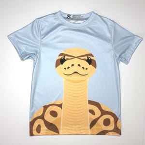May include: A light blue short-sleeve t-shirt featuring a large graphic of a yellow and brown snake with a patterned body. The shirt has a size S tag at the neckline with text that reads 100% Polyester, Machine wash cold, Tumble dry low.