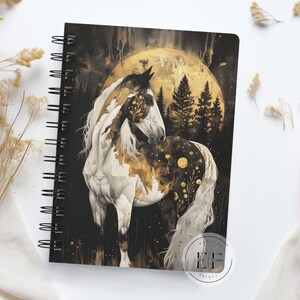 May include: A black and gold spiral notebook with a white horse illustration. The horse is standing in front of a full moon and trees. The text "Manifest Your Dreams" is printed on the bottom right corner of the notebook.