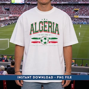 以下が含まれることがあります： 胸元に緑と赤のALGERIAの文字、EST. 1962、サッカーボール、そしてかすれた筆跡スタイルのLES FENNECSとLES GUERRIERS DU SAHARAの文字がプリントされた、白い半袖Tシャツ。