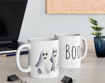 May include: Two white ceramic mugs. One mug features a watercolor illustration of two friendly ghosts. The other mug has the word "BOO" written in black.