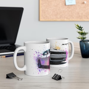 May include: Two white ceramic mugs with a car design. One mug features a side view of a white car with purple and pink paint splatter. The other mug shows the front of a gray car with the text "SRT" on the side.