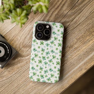 May include: A white phone case with a pattern of green and light blue watercolor shamrocks. The case is on a wooden surface, with a camera and plant in the background.