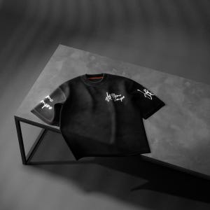 May include: Black short-sleeved t-shirt with white script on the sleeves and chest. The shirt has a crew neck with a thin orange accent. The garment is displayed on a dark gray surface.