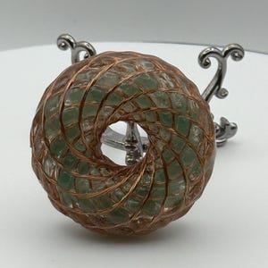 May include: A green stone donut-shaped pendant wrapped in copper wire, displayed on a silver metal stand. The pendant has a hole in the centre and is made of small, irregular green stones. The copper wire is intricately woven around the stone.