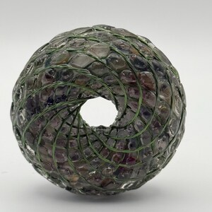 May include: A decorative, hollow sphere made from small, irregularly shaped, translucent stones, bound with green wire. The stones have a mix of purple, brown, and clear tones. The sphere has a central opening.