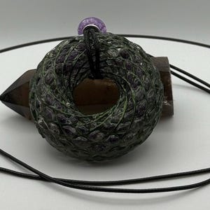 May include: A dark green and black pendant necklace with a circular, textured design. The pendant has a hole in the centre and is strung on a black cord. A purple bead is at the top of the pendant.