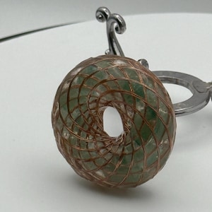 May include: A handcrafted, donut-shaped pendant featuring a green stone encased in a copper wire cage. The pendant is suspended from a silver-coloured metal bail, creating a unique and artistic jewellery piece.