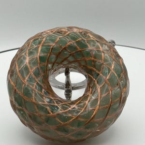 May include: A green, toroidal object with a copper wire grid pattern. The object is displayed on a silver stand against a white background. The design features a central hole and a textured surface.