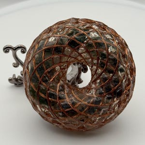 May include: A decorative, donut-shaped object with a copper wire grid over a dark interior. A silver-coloured, ornate handle extends from the side. The object is likely a unique home decor item or sculpture.