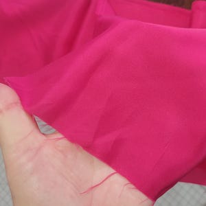 Peut inclure: Un échantillon de tissu fuchsia vif, présenté pour montrer sa texture et sa couleur. Le tissu semble doux et léger, avec un léger éclat. Les bords sont bruts, indiquant qu'il pourrait s'agir d'une pièce coupée ou d'un échantillon de matériau.