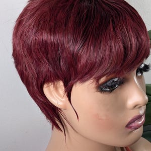 May include: A short, burgundy wig with a layered pixie cut. The wig is styled with a side parting and has a natural-looking hairline. The hair is a deep, rich shade of red.