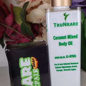 Peut inclure: Une bouteille blanche d'"Huile Corporelle Mixte à la Noix de Coco" avec un bouchon doré et une boîte noire de "Rare Repair Soap" sont présentées. La boîte de savon a du texte vert et orange. Le fond présente des fleurs colorées.