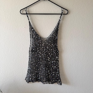 May include: A black sequined tank top with thin straps hangs on a black hanger against a white wall. The top is covered in small, shiny sequins, creating a shimmering effect. The neckline is a deep V-shape.