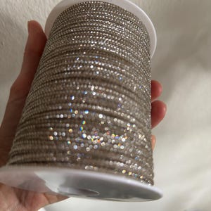 May include: A reel of silver rhinestone trim. The trim is wound around a white plastic reel. The rhinestones sparkle and reflect light, creating a shimmering effect. Suitable for craft and design projects. The reel is held by a hand.