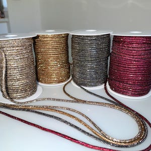 May include: Four spools of metallic cord in silver, gold, dark gray, and deep red. Each spool is wrapped with a continuous length of shimmering, textured cord that extends outward, displaying the reflective, glittery surface of the material.