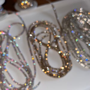 May include: Multiple spools of silver-toned rhinestone chain are coiled on a white surface. The chains feature small, reflective crystals that create a shimmering, multi-colored bokeh effect, highlighting the metallic texture and light-catching quality.