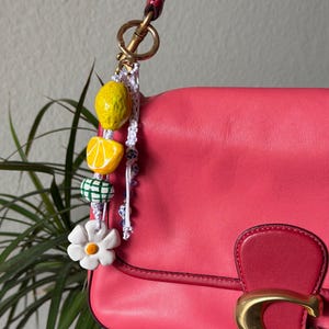 May include: A pink leather handbag with a gold clasp and a decorative charm. The charm features a yellow lemon, a lemon slice, a green and white checkered bead, and a white flower with a yellow centre. The charm is attached to the bag's strap.