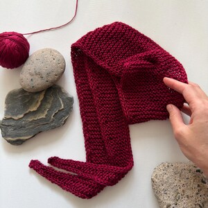 May include: A knitted burgundy hat with a long scarf-like extension. The hat and scarf are made of a thick yarn, with a visible knit texture. A ball of matching yarn and several stones are arranged nearby.