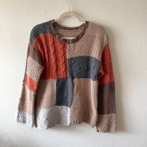 Könnte beinhalten: Ein langärmeliger Pullover mit Patchwork-Design und verschiedenen Strickmustern wie Zopfmuster, Noppen und Perlmuster. Das Kleidungsstück besteht aus rechteckigen Bahnen in den Farben gebranntes Orange, Anthrazit, Beige und Hellbraun.