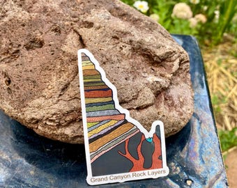 May include: A sticker featuring a colorful illustration of Grand Canyon rock layers. The sticker has a white border and the text "Grand Canyon Rock Layers" at the bottom. The sticker is placed on a dark blue surface next to a large, textured rock.