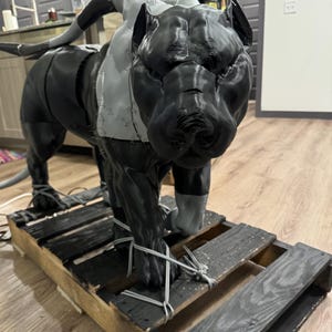 May include: A large, 3D-printed sculpture of a dog, primarily black with gray accents, is secured to a wooden pallet with rope. The dog is in a walking pose, with detailed features. The sculpture appears to be in the process of being assembled.