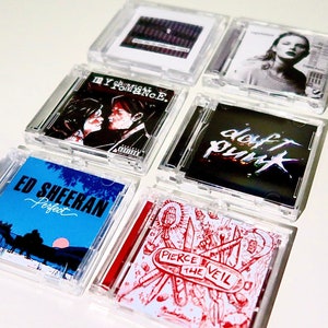 Puede incluir: Seis estuches de CD diferentes con diversas portadas de álbum. Las portadas presentan una variedad de artistas y géneros, incluyendo pop, rock y música electrónica. Los estuches son de plástico transparente y muestran claramente el arte del álbum.