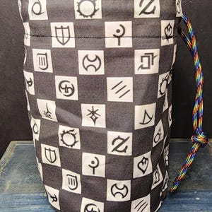 May include: A cylindrical fabric drawstring pouch featuring a repeating checkerboard pattern of black and white squares. Each square contains a unique black graphic symbol. The pouch is cinched with a braided, multi-colored rainbow cord.