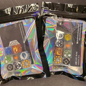 Puede incluir: Dos bolsas hologr&aacute;ficas que contienen juegos de dados personalizados. Cada bolsa incluye varios dados cuadrados con diversos iconos gr&aacute;ficos coloridos, como s&iacute;mbolos de &aacute;rboles, soles y formas abstractas. Dentro hay una tarjeta negra con el texto blanco AM Lit Arts.