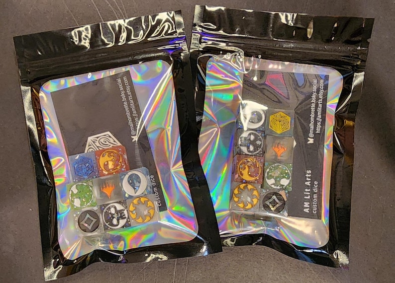 Puede incluir: Dos bolsas hologr&aacute;ficas que contienen juegos de dados personalizados. Cada bolsa incluye varios dados cuadrados con diversos iconos gr&aacute;ficos coloridos, como s&iacute;mbolos de &aacute;rboles, soles y formas abstractas. Dentro hay una tarjeta negra con el texto blanco AM Lit Arts.