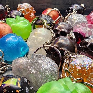 May include: A collection of colorful skull-shaped keychains in various colors, including red, blue, green, and black. Some keychains have glitter or transparent designs. Each keychain has a silver chain.