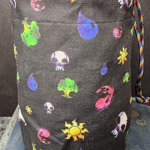 May include: A cylindrical fabric drawstring bag in black, featuring a repeating pattern of colorful icons including yellow suns, blue water droplets, green trees, pink flames, and white skulls. The bag includes a rainbow-colored cord drawstring.