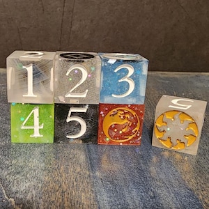 Puede incluir: Un conjunto de seis dados cuadrados de resina en varios colores, incluyendo transparente, gris, azul, verde, negro y rojo. Los dados muestran n&uacute;meros blancos del uno al cinco y un s&iacute;mbolo de sol dorado. Cada cubo tiene un acabado liso con purpurina.