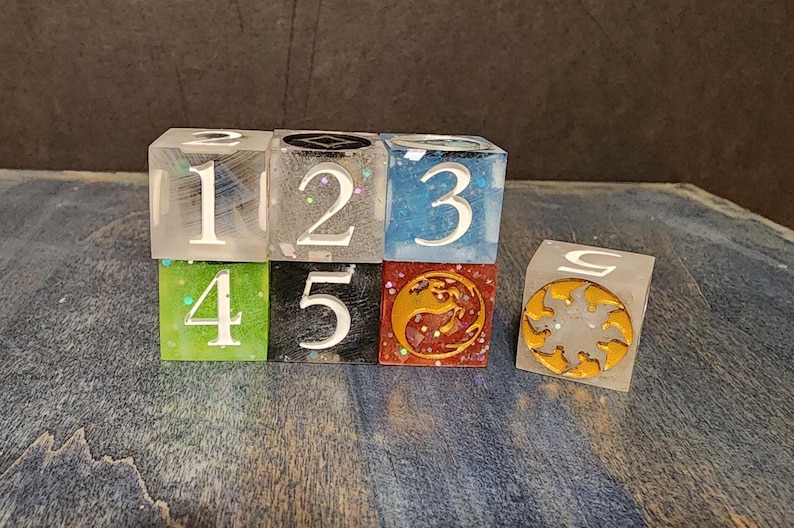 Puede incluir: Un conjunto de seis dados cuadrados de resina en varios colores, incluyendo transparente, gris, azul, verde, negro y rojo. Los dados muestran n&uacute;meros blancos del uno al cinco y un s&iacute;mbolo de sol dorado. Cada cubo tiene un acabado liso con purpurina.