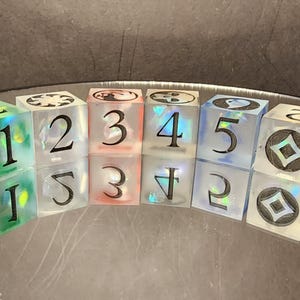 Könnte beinhalten: Set aus sechs durchsichtigen Harzwürfeln mit schwarzen Zahlen und Symbolen. Die Würfel sind in einer gebogenen Linie angeordnet, wobei jeder Würfel eine andere Zahl von 1 bis 5 und ein Symbol anzeigt. Die Würfel haben einen holografischen Effekt.