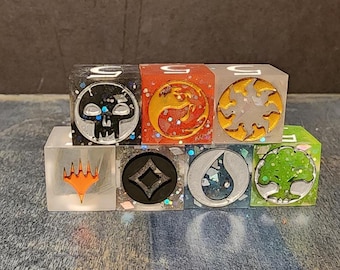 Puede incluir: Un conjunto de siete dados de resina translúcida con símbolos de maná de Magic: The Gathering. Los dados incluyen colores negro, rojo, blanco, transparente, gris oscuro, azul y verde con purpurina incrustada y símbolos metálicos.
