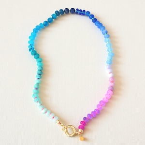 May include: A beaded necklace with a gradient of colors, transitioning from dark blue to light blue, then to pink and purple. The necklace has a gold clasp and a small gold charm. The beads appear to be small and round.