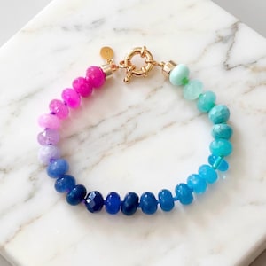 Gradient gemstone bracelet featuring genuine pink, lavender, cobalt blue, and aqua blue faceted beads with 14k gold vermeil accents and a gold toggle clasp on a white marble background.