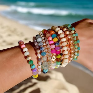 May include: A stack of colorful beaded bracelets with a gold charm in the center. The bracelets are made of various materials, including glass, wood, and metal. The bracelets are worn on a person's wrist.