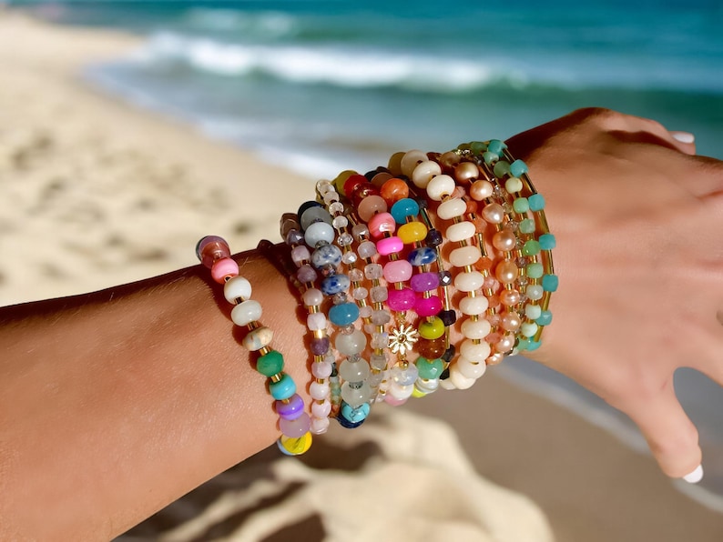 May include: A stack of colorful beaded bracelets with a gold charm in the center. The bracelets are made of various materials, including glass, wood, and metal. The bracelets are worn on a person's wrist.
