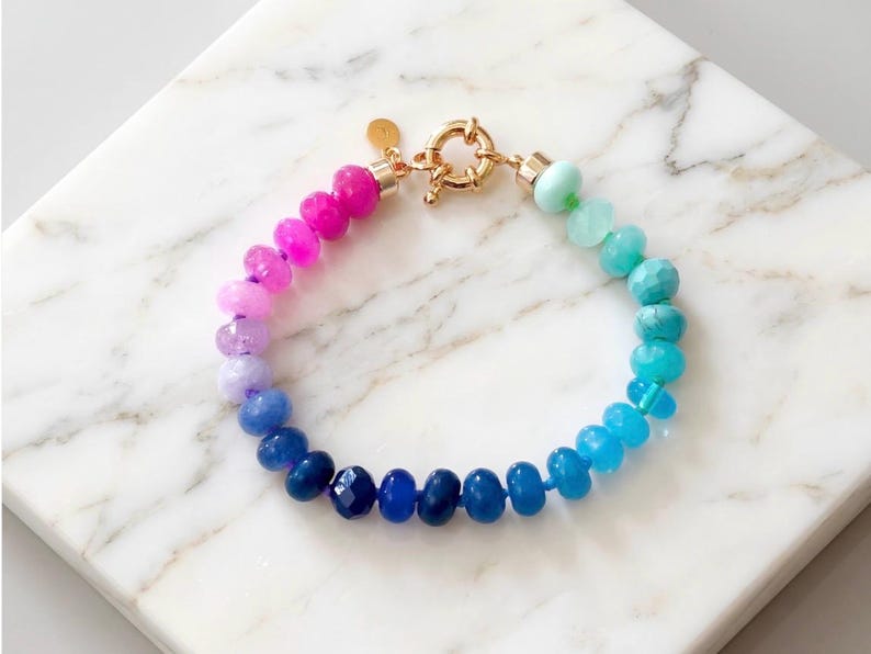 Gradient gemstone bracelet featuring genuine pink, lavender, cobalt blue, and aqua blue faceted beads with 14k gold vermeil accents and a gold toggle clasp on a white marble background.