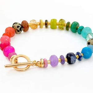 Puede incluir: Una colorida pulsera de cuentas con un cierre dorado. La pulsera presenta una variedad de cuentas de piedras preciosas en tonos de rojo, rosa, naranja, amarillo, verde, azul y morado. Las cuentas son de varias formas y tamaños.