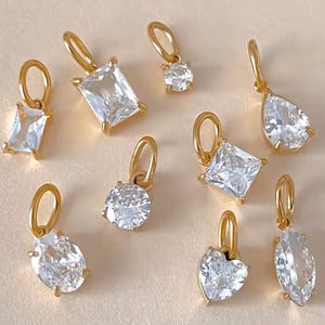 Gold diamond crystal charms displayed on a white background, each featuring a unique rhinestone cut — pear, heart, round, oval, marquise, princess, emerald, and radiant. The clear faceted stones shimmer in the light, framed in gold settings with looped bails for attaching to necklaces or bracelets. Elegant minimalist photo showing jewelry-making pendants available in both gold and silver options.  Perfect for diy jewelry to use as add-on charms for any charm necklace or charm bracelet.