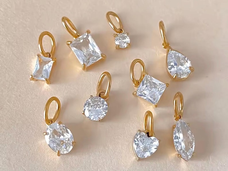 Gold diamond crystal charms displayed on a white background, each featuring a unique rhinestone cut — pear, heart, round, oval, marquise, princess, emerald, and radiant. The clear faceted stones shimmer in the light, framed in gold settings with looped bails for attaching to necklaces or bracelets. Elegant minimalist photo showing jewelry-making pendants available in both gold and silver options.  Perfect for diy jewelry to use as add-on charms for any charm necklace or charm bracelet.