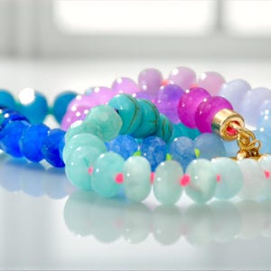 Hand-knotted gemstone necklace featuring turquoise, royal blue, and lilac beads with a gold clasp, laid out in a soft ombré gradient.