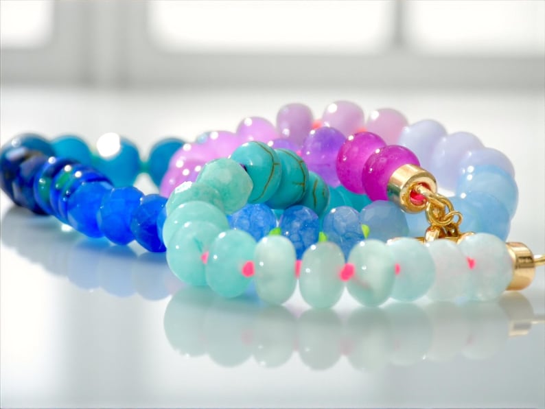 Hand-knotted gemstone necklace featuring turquoise, royal blue, and lilac beads with a gold clasp, laid out in a soft ombré gradient.