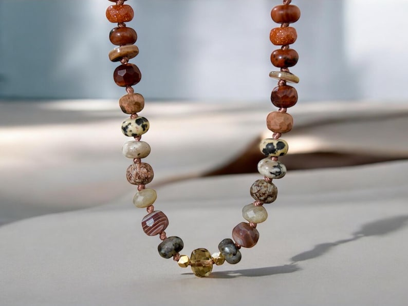 A long necklace made of various colored stones. The stones are in shades of brown, creams, white, and natural hues. The necklace has a gold clasp.
