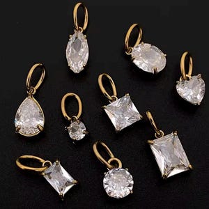 Gold diamond crystal charms displayed on a black background, each featuring a unique rhinestone cut — pear, heart, round, oval, marquise, princess, emerald, and radiant. The clear faceted stones shimmer in the light, framed in gold settings with looped bails for attaching to necklaces or bracelets. Elegant minimalist photo showing jewelry-making pendants available in both gold and silver options.  Perfect for diy jewelry to use as add-on charms for any charm necklace or charm bracelet.