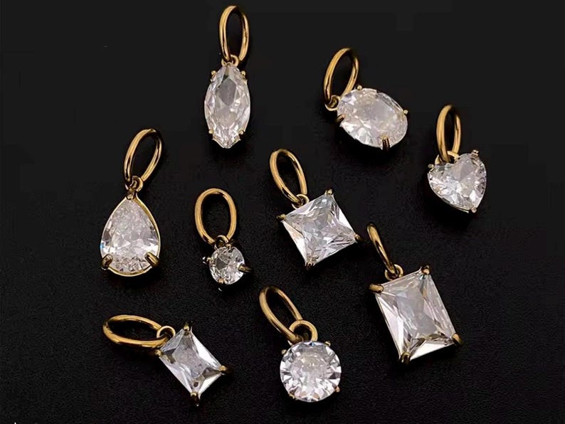 Gold diamond crystal charms displayed on a black background, each featuring a unique rhinestone cut — pear, heart, round, oval, marquise, princess, emerald, and radiant. The clear faceted stones shimmer in the light, framed in gold settings with looped bails for attaching to necklaces or bracelets. Elegant minimalist photo showing jewelry-making pendants available in both gold and silver options.  Perfect for diy jewelry to use as add-on charms for any charm necklace or charm bracelet.