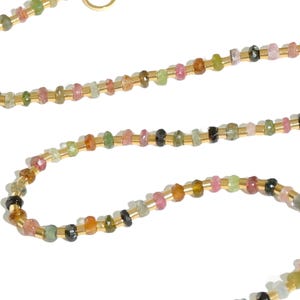 May include: A delicate gold chain necklace with small, multi-colored gemstones. The necklace features a variety of colors, including pink, green, and black.